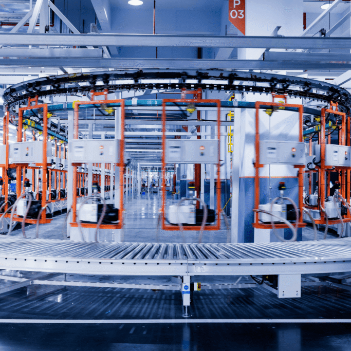 Circular overhead conveyor with orange frames in a spacious industrial facility; two workers in orange helmets sit at left with blue crates nearby.
